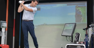 A man in a white polo shirt and blue dress pants swings a golf club at a ball in the swing bay of an indoor simulator.