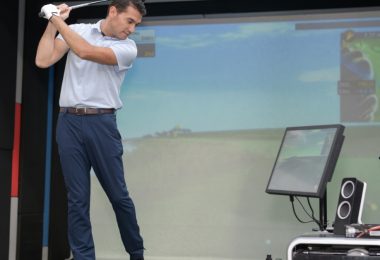 A man in a white polo shirt and blue dress pants swings a golf club at a ball in the swing bay of an indoor simulator.