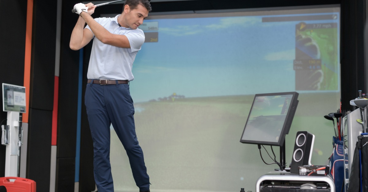 A man in a white polo shirt and blue dress pants swings a golf club at a ball in the swing bay of an indoor simulator.