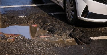A white car driving past a large pothole in the asphalt. The pothole contains water and multiple bumps.