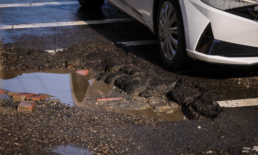 A white car driving past a large pothole in the asphalt. The pothole contains water and multiple bumps.