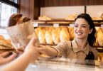 A woman standing behind the counter of a bakery reaches over to hand a pastry in a paper bag to awaiting hands.