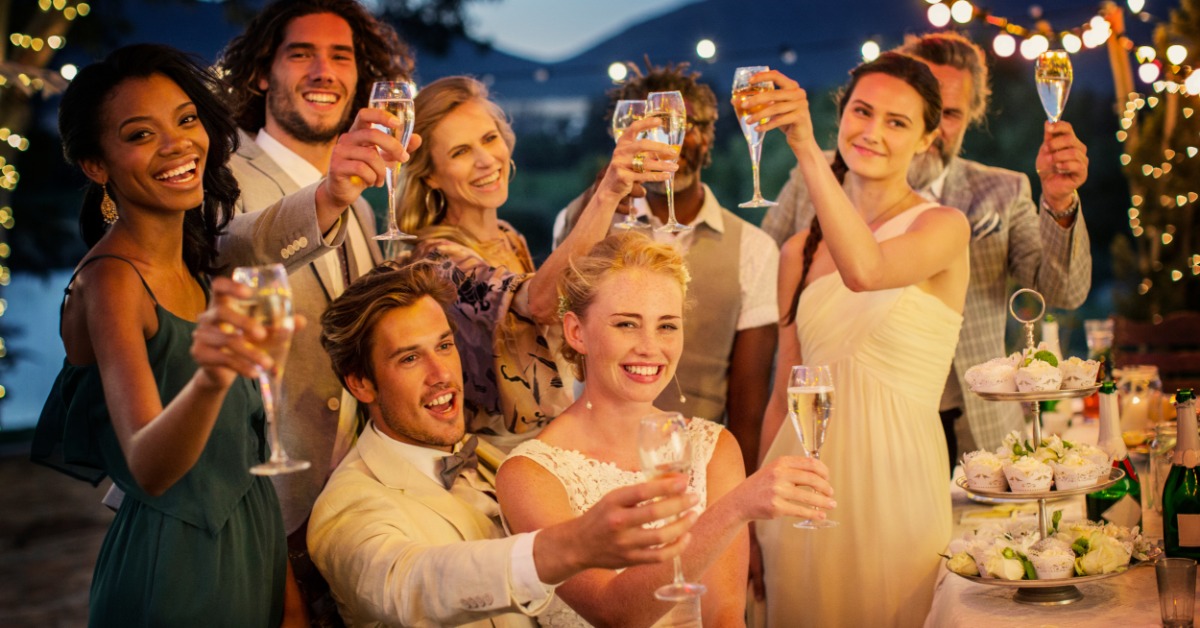 A group of people are standing around a sitting couple. The group is outside and the sun is down.