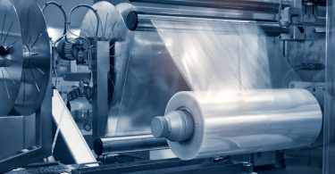 A plastic film roll feeding clear material through metal rollers on an automated machine under blue light.