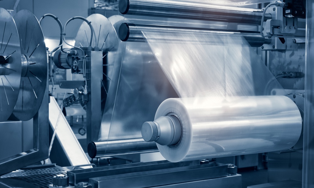 A plastic film roll feeding clear material through metal rollers on an automated machine under blue light.