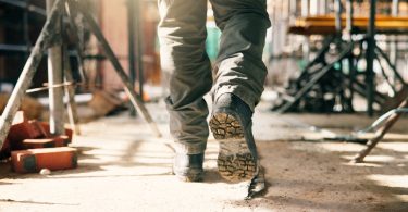Low-angle view of a person's boots as they walk through a construction site. Bricks and scaffolding are visible.
