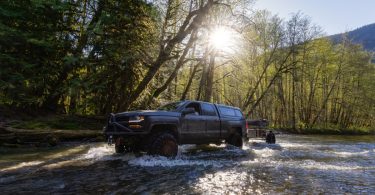 A pickup truck with a large metal protection bar in the front and a large bed cover crossing through a river.