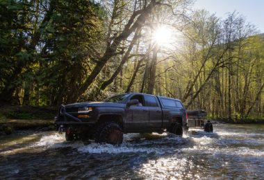 A pickup truck with a large metal protection bar in the front and a large bed cover crossing through a river.