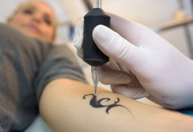 A close-up of a hand wearing a white latex glove, holding an ink pen while tattooing a small shape on a woman's arm.