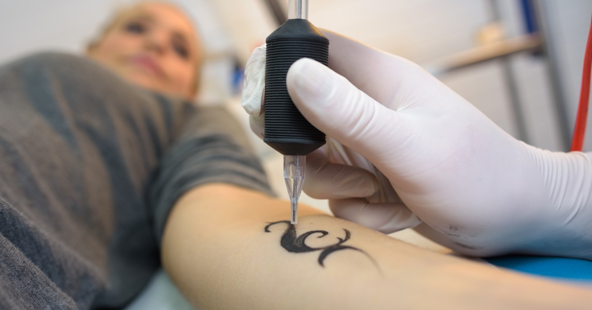 A close-up of a hand wearing a white latex glove, holding an ink pen while tattooing a small shape on a woman's arm.