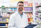 A smiling medical professional in a white coat holds a clipboard between pharmacy shelves stocked with medicine.