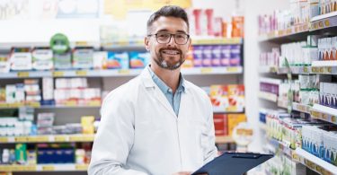 A smiling medical professional in a white coat holds a clipboard between pharmacy shelves stocked with medicine.