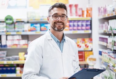 A smiling medical professional in a white coat holds a clipboard between pharmacy shelves stocked with medicine.