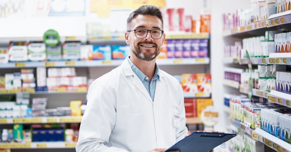 A smiling medical professional in a white coat holds a clipboard between pharmacy shelves stocked with medicine.