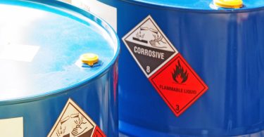 A close-up of two blue drums with a "corrosive" and "flammable liquid" label on the side. Sun reflects off the top of the drums.