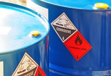 A close-up of two blue drums with a "corrosive" and "flammable liquid" label on the side. Sun reflects off the top of the drums.