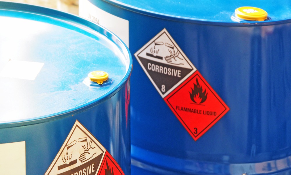 A close-up of two blue drums with a "corrosive" and "flammable liquid" label on the side. Sun reflects off the top of the drums. A close-up of two blue drums with a "corrosive" and "flammable liquid" label on the side. Sun reflects off the top of the drums.