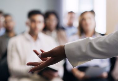 A professional speaking in front of a crowd at a trade show. The speaker extends their left arm to the left.