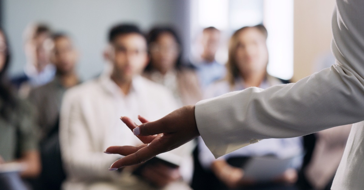 A professional speaking in front of a crowd at a trade show. The speaker extends their left arm to the left.
