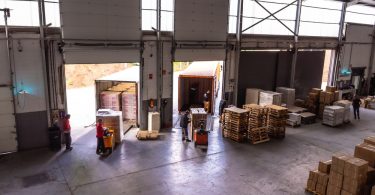 Workers and forklifts move goods near loading bays inside a warehouse with pallets and packaged inventory.