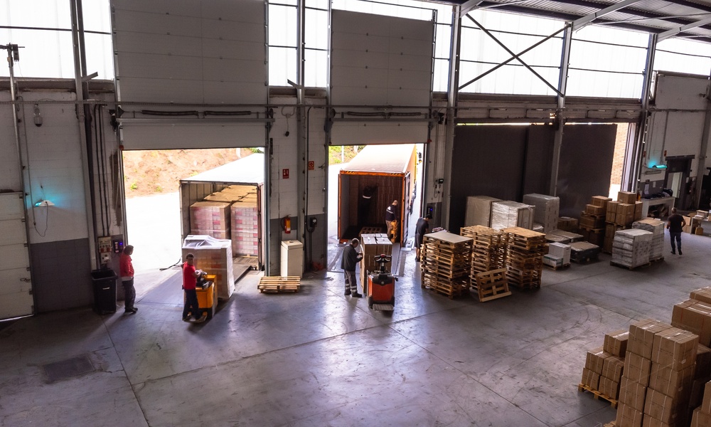 Workers and forklifts move goods near loading bays inside a warehouse with pallets and packaged inventory.