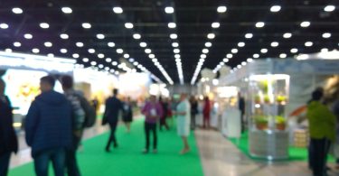 Blurred people walking through a bright trade show hall with booth displays, glossy floors, and ceiling lights.