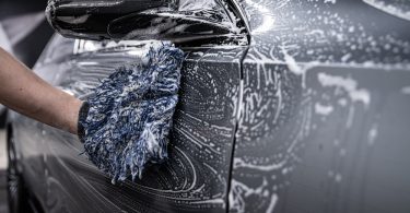 A gloved hand applying coating to a glossy black car panel near a mirror with a small applicator pad.