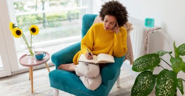 A woman with curly hair journals in a teal armchair beside a sunny window with sunflowers and a cup of tea.