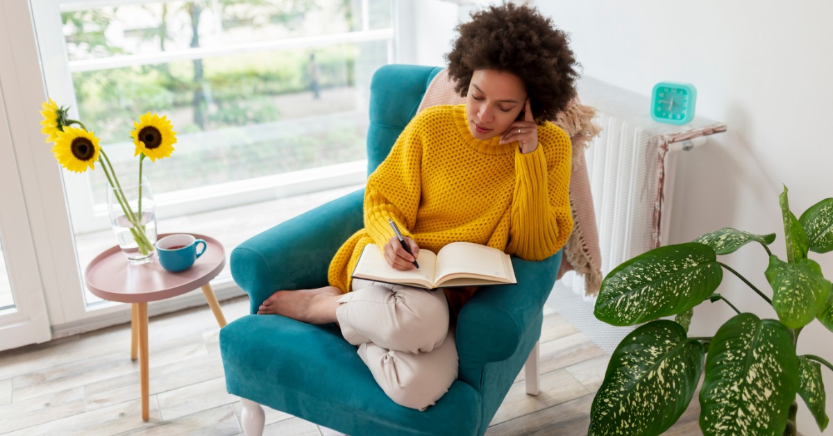 A woman with curly hair journals in a teal armchair beside a sunny window with sunflowers and a cup of tea.
