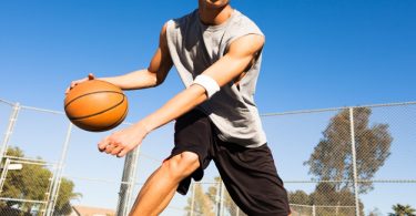A young person dribbling a basketball on an outdoor court with a chain-link fence and blue sky behind them.