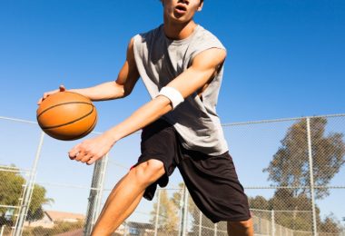 A young person dribbling a basketball on an outdoor court with a chain-link fence and blue sky behind them.