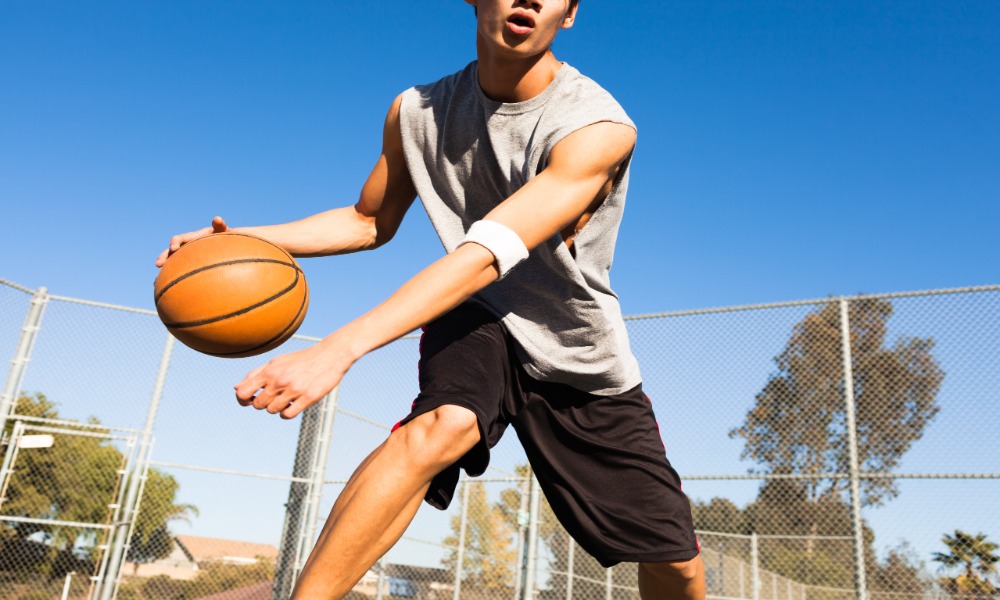 A young person dribbling a basketball on an outdoor court with a chain-link fence and blue sky behind them. A young person dribbling a basketball on an outdoor court with a chain-link fence and blue sky behind them.