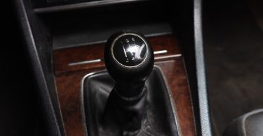 A close-up of a manual transmission gear shift knob inside a car. The knob shows five speeds and has leather around the stem.