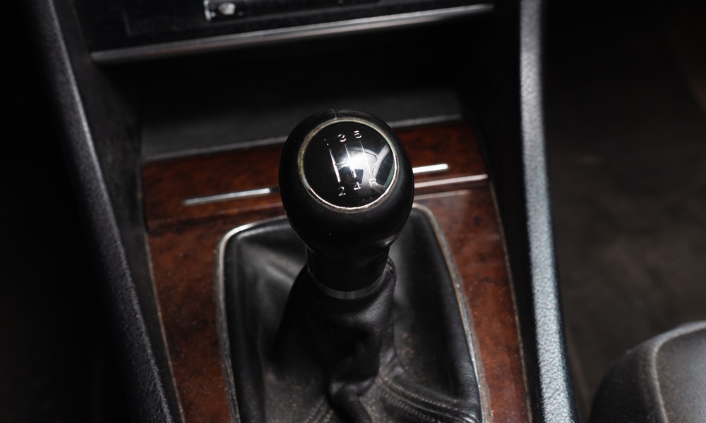 A close-up of a manual transmission gear shift knob inside a car. The knob shows five speeds and has leather around the stem.