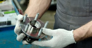 Someone wearing white fabric gloves holds a metal auto part in their hand. There is a worn blue table beneath the part.