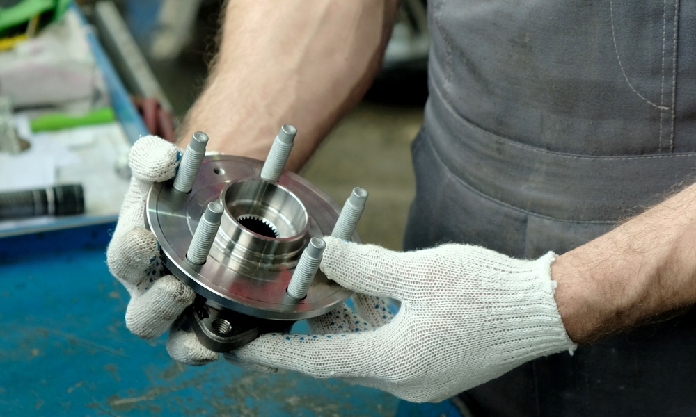 Someone wearing white fabric gloves holds a metal auto part in their hand. There is a worn blue table beneath the part.