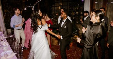 A bride and groom surrounded by a lively dancing crowd during a wedding. The couple dances next to a table.