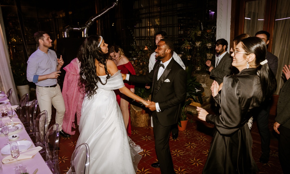 A bride and groom surrounded by a lively dancing crowd during a wedding. The couple dances next to a table.