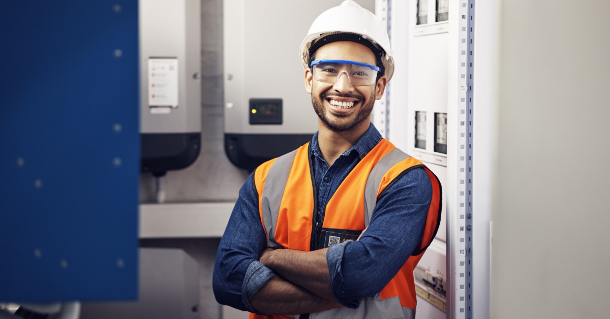 An electrician crossing their arms and smiling next to a wall full of electrical panels in a workplace.