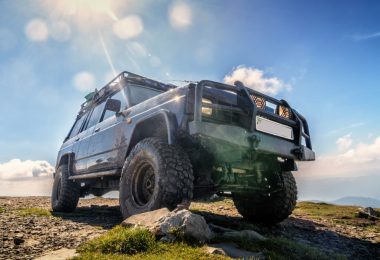 A lifted off-road SUV with mud tires and a roof rack parked on a rocky mountain summit under the bright sun.