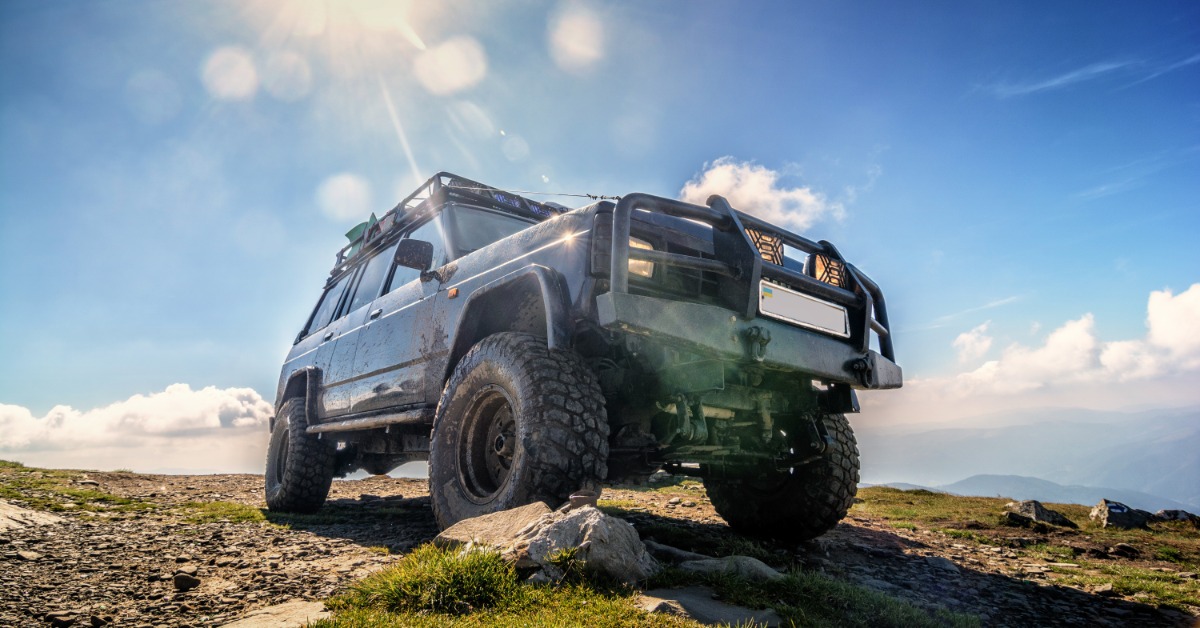 A lifted off-road SUV with mud tires and a roof rack parked on a rocky mountain summit under the bright sun.