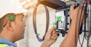 A fiber network engineer wears a white hard hat at the fiber panel attached to the exterior of a property.