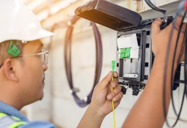 A fiber network engineer wears a white hard hat at the fiber panel attached to the exterior of a property.