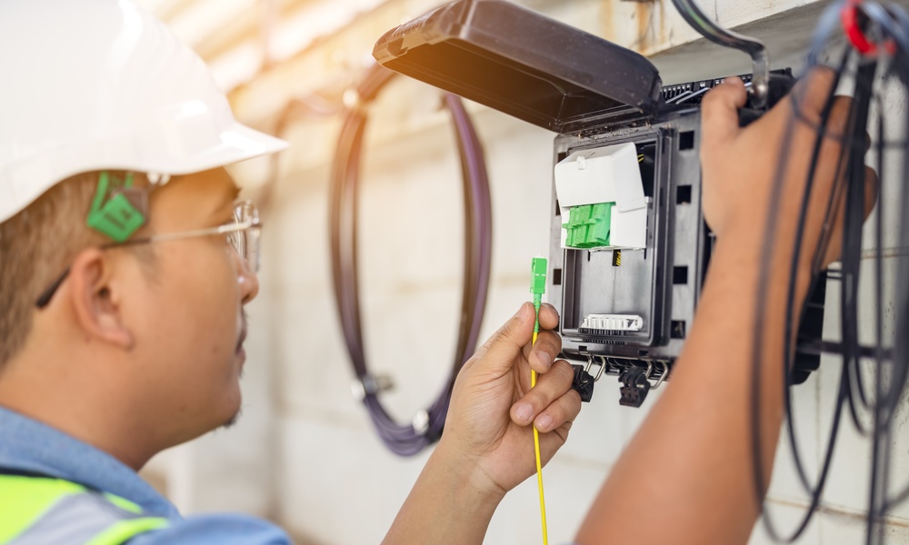 A fiber network engineer wears a white hard hat at the fiber panel attached to the exterior of a property.