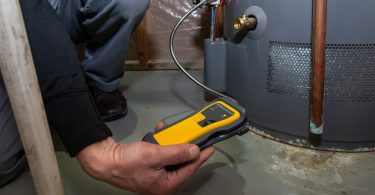 Technician holds a yellow testing device beside a water heater, checking for potential gas or equipment hazards.