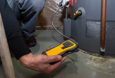 Technician holds a yellow testing device beside a water heater, checking for potential gas or equipment hazards.