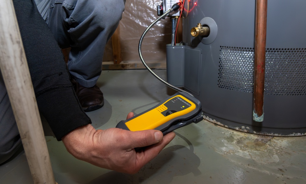 Technician holds a yellow testing device beside a water heater, checking for potential gas or equipment hazards.