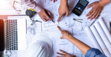Several people pointing at blueprints on a desk with a laptop, calculator, hard hat, rolled papers, and tools nearby.