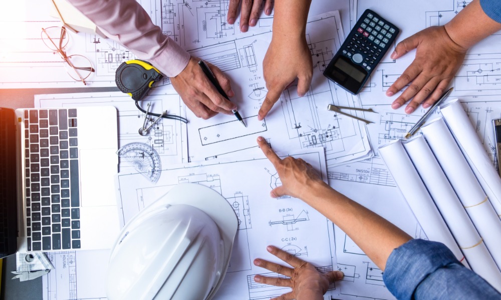 Several people pointing at blueprints on a desk with a laptop, calculator, hard hat, rolled papers, and tools nearby.