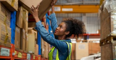 A worker wearing a yellow safety vest lifting a cardboard box from a shelf in a warehouse aisle with multiple racks.